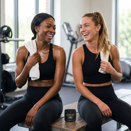 Two women laughing on a gym bench after a workout, CHARGE MELT tin between them